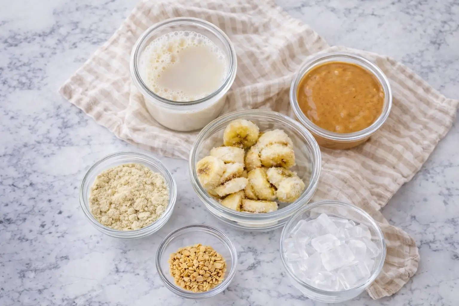 Ingredients for peanut butter banana smoothie including banana slices, peanut butter, milk, ice, and protein powder on marble surface