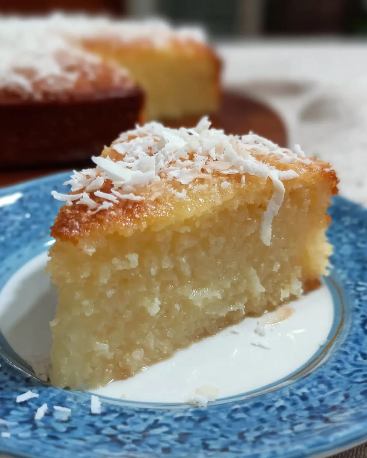 Slice of coconut magic cake dusted with powdered sugar on a white plate with a whole coconut in the background. Coconut Cake with Condensed Milk